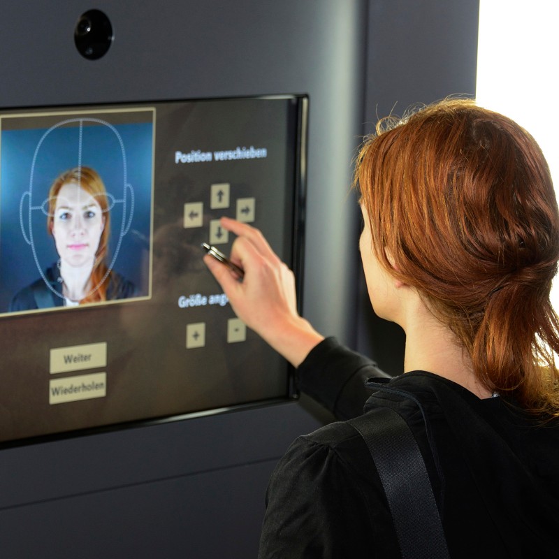 Frau
                                 mit rötlichen Haaren von hinten, bedient einen großen Touchscreen mit Porträtfoto und Steuertasten für Position und Größe.
                                 (KI-generierter Alt-Text, erstellt mit GPT-4.1-mini)