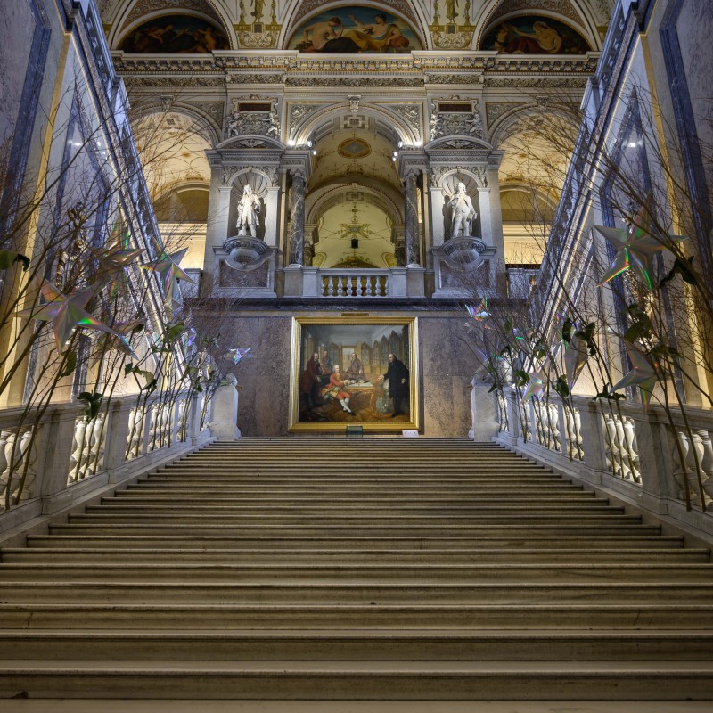Wide marble staircase in an ornate foyer, decorated on both sides with branches and Christmas stars; large painting
                                 and statues in the background. (AI-generated alt-text, generated with GPT-4.1-mini)