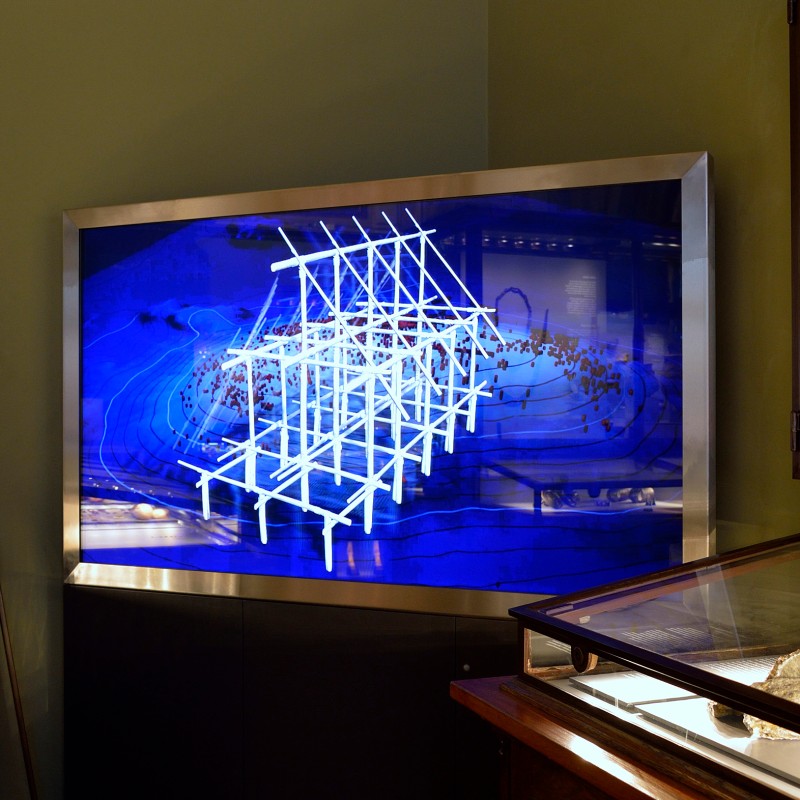 Modell
                                 eines hölzernen Pfahlbaus aus weißen Stäben vor blauem Hintergrund. Das Modell steht in einer beleuchteten Vitrine. (KI-generierter
                                 Alt-Text, erstellt mit GPT-4.1-mini)