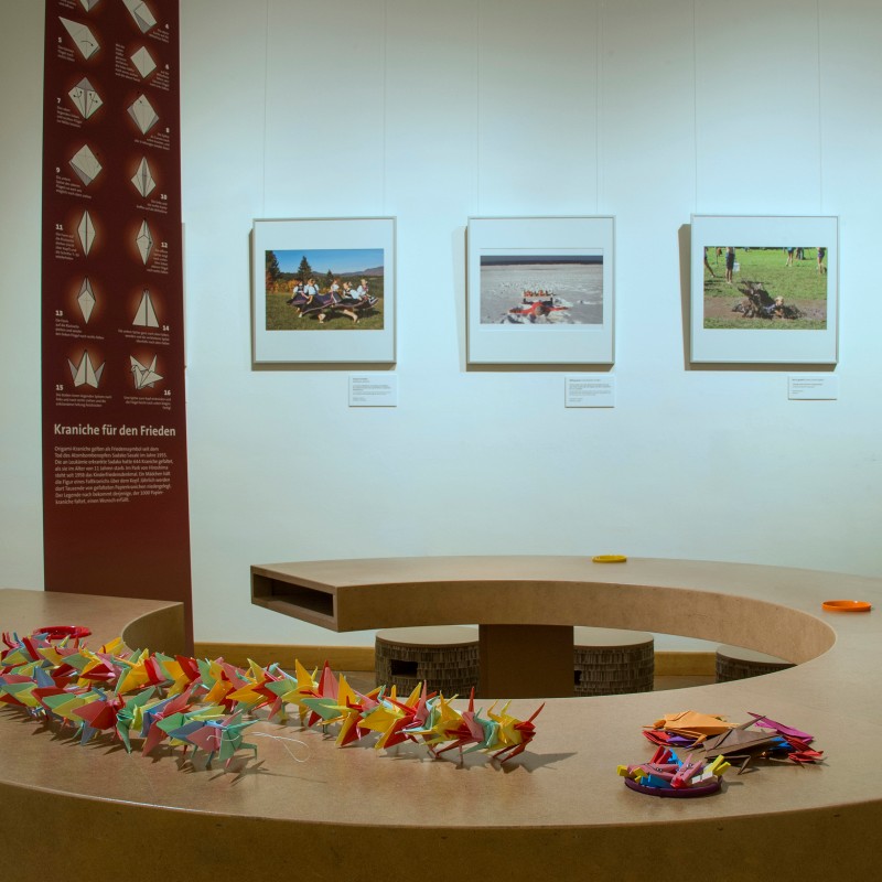 Multiple colorful paper cranes rest on a round table. Three photographs and a folding instruction diagram hang on
                                 the wall behind. (AI-generated alt-text, generated with GPT-4.1-mini)