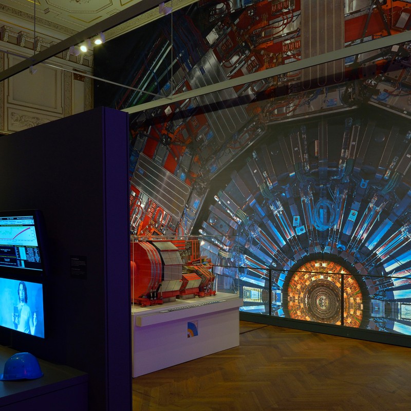 Große
                                 Projektion eines Teilchendetektors mit rundem, komplexem Aufbau im Ausstellungsraum, daneben Wand mit Monitoren und zwei Schutzhelmen
                                 auf einem Tisch. (KI-generierter Alt-Text, erstellt mit GPT-4.1-mini)