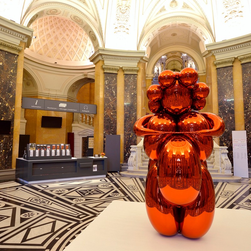 Reflective orange sculpture of the Venus of Willendorf in an ornate hall with marble columns and black-and-white
                                 tile floor. (AI-generated alt-text, generated with GPT-4.1-mini)