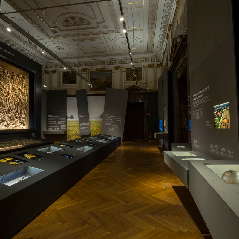 Dimly lit exhibition room with illuminated display cases, wall texts, and a large relief with many figures on the
                                 wall. (AI-generated alt-text, generated with GPT-4.1-mini)