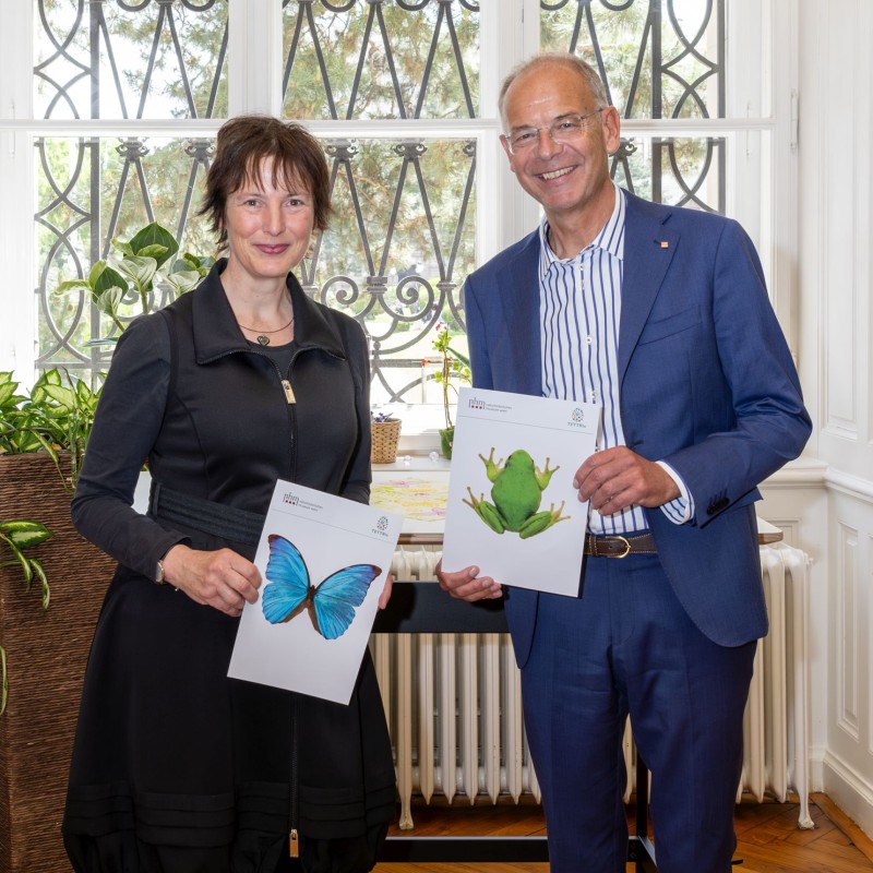 A woman dressed in black holding an A4 sheet of paper with a blue butterfly on it, standing next to a man dressed in a blue suit holding an A4 sheet of paper with a green frog on it.