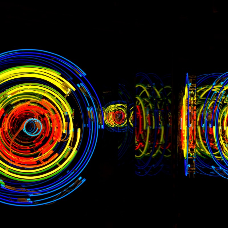 Kreisförmige Lichtinstallation mit roten, gelben und blauen Neonröhren, mehrfach in einem dunklen Raum gespiegelt.
                                 (KI-generierter Alt-Text, erstellt mit GPT-4.1-mini)