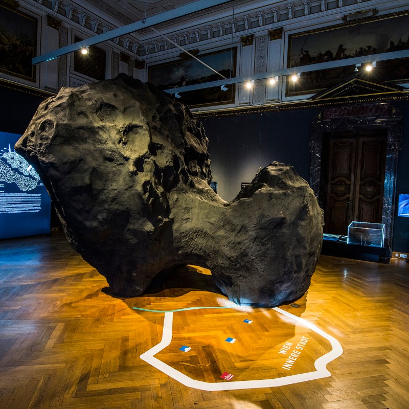 Großes, unregelmäßig geformtes Objekt auf einem Museumsparkett, in Ausstellungsraum mit Infotafeln und Leuchtmarkierung
                                 auf dem Boden. (KI-generierter Alt-Text, erstellt mit GPT-4.1-mini)