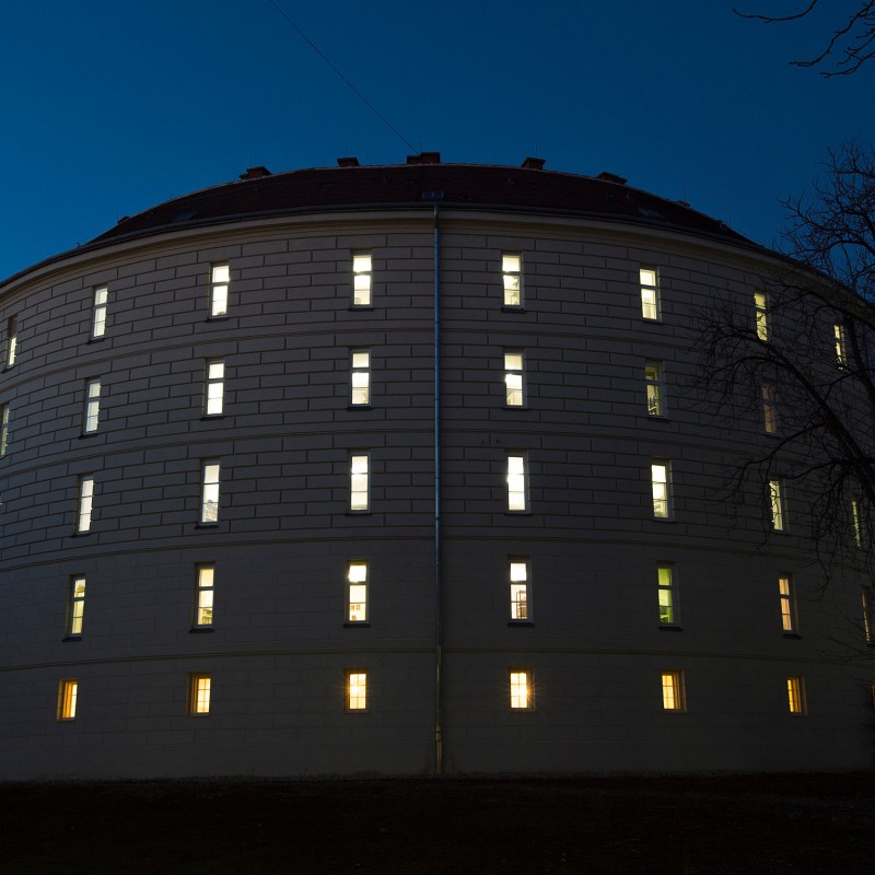 Großes, rundes Gebäude mit erleuchteten Fenstern in der Dämmerung, kahle Bäume im Vordergrund. (KI-generierter Alt-Text,
                                 erstellt mit GPT-4.1-mini)