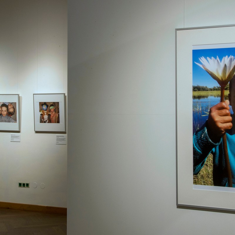 Gallery exhibition with photographs on white walls, foreground features framed photo of a child holding a white
                                 flower by a body of water. (AI-generated alt-text, generated with GPT-4.1-mini)
