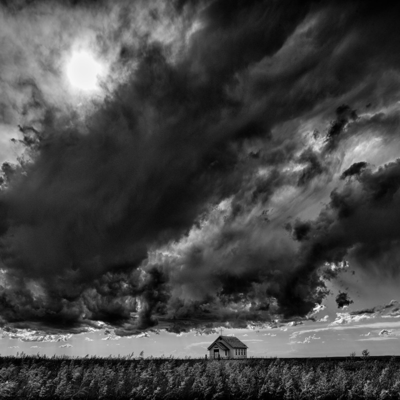 Small church in an open landscape under a dramatic cloudy sky with a faintly visible sun. (AI-generated alt-text,
                                 generated with GPT-4.1-mini)