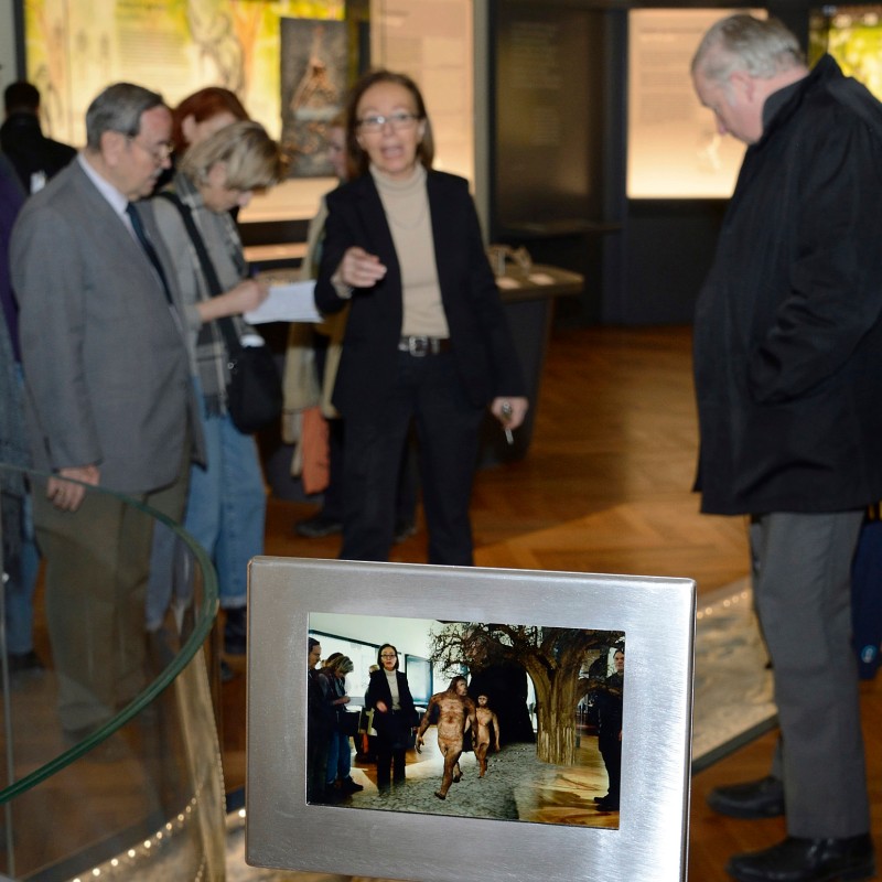 Mehrere Erwachsene stehen in einer Museumsausstellung und betrachten Exponate. Im Vordergrund befindet sich ein
                                 Schild mit einem Foto, das eine ähnliche Szene zeigt. (KI-generierter Alt-Text, erstellt mit GPT-4.1-mini)