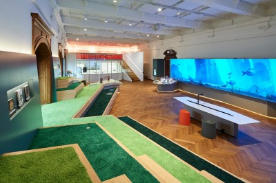 Deck 50: Innenraum von Deck 50 mit grünen Teppichflächen, hölzernem Boden, stufenförmigen Sitzgelegenheiten und großer digitaler Wand mit blauer Unterwasserszenerie. (KI-generierter Alt-Text, erstellt mit GPT-4.1-mini)