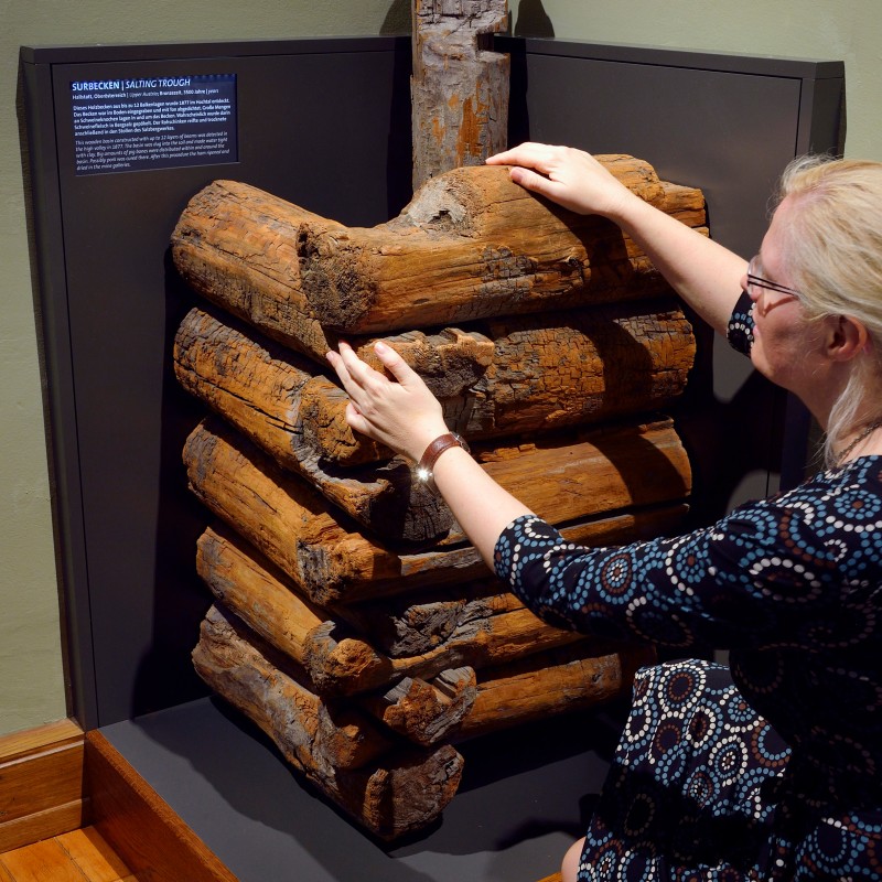 Frau mit Brille tastet ein aus mehreren Holzstücken zusammengesetztes Objekt im Ausstellungsraum ab. (KI-generierter
                                 Alt-Text, erstellt mit GPT-4.1-mini)