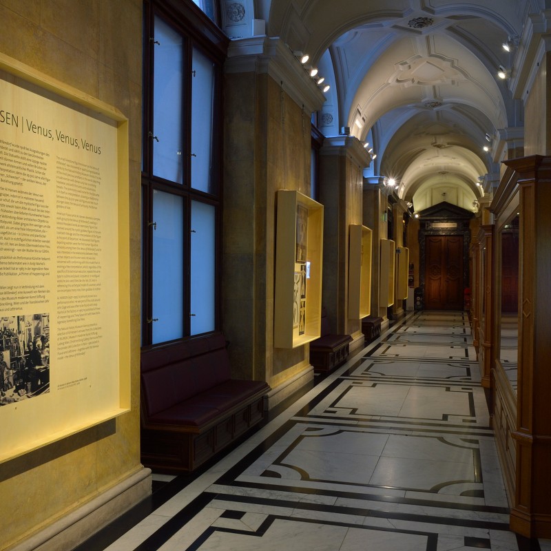 Long museum corridor with exhibition wall text “Al Hansen | Venus Venus Venus”, illuminated panels and seating alcoves.
                                 (AI-generated alt-text, generated with GPT-4.1-mini)