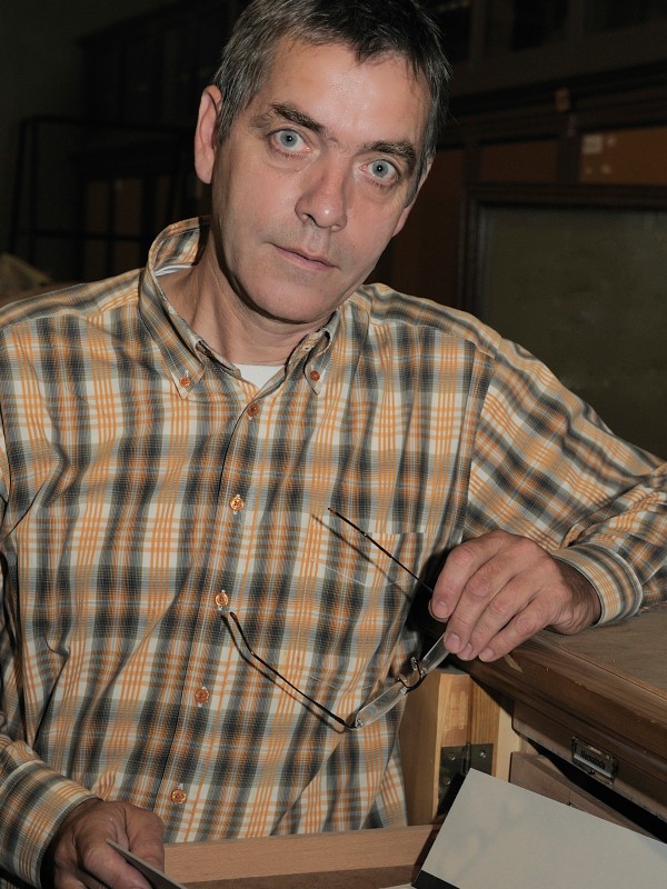 Mann mit kariertem
                                 Hemd hält eine Brille in der rechten Hand und öffnet eine Schublade mit Blattpapieren. (KI-generierter Alt-Text, erstellt
                                 mit GPT-4.1-mini)