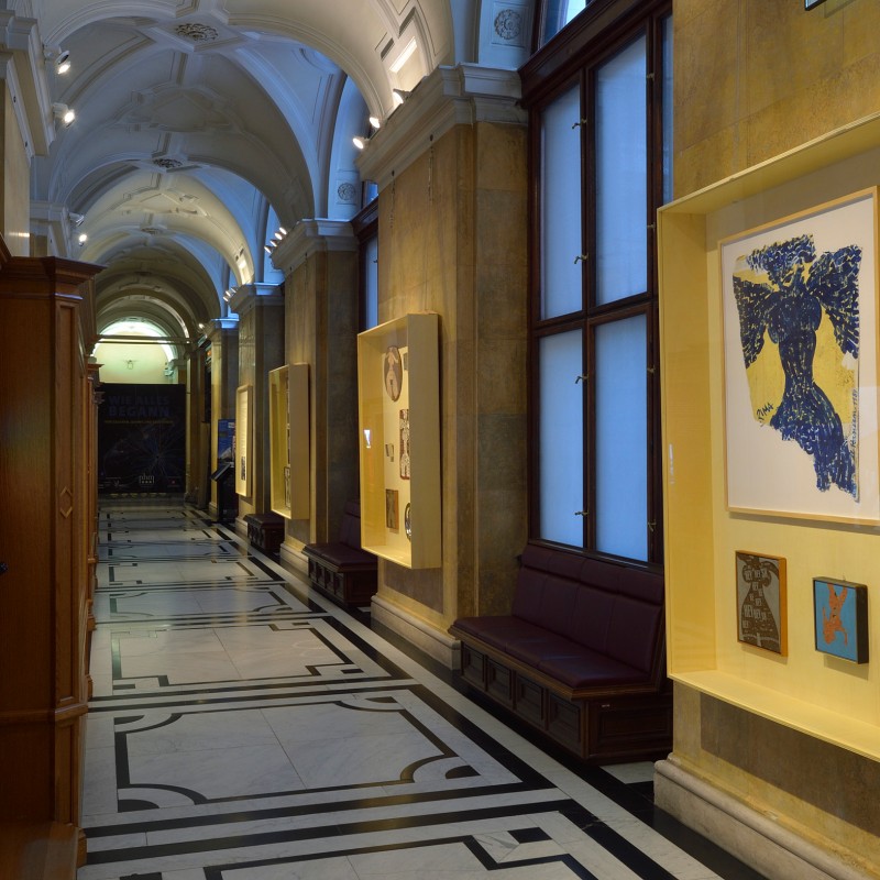 Museum corridor with illuminated display cases on the wall, featuring modern graphic art and collages. Daylight
                                 streams through large windows, benches along the side. (AI-generated alt-text, generated with GPT-4.1-mini)