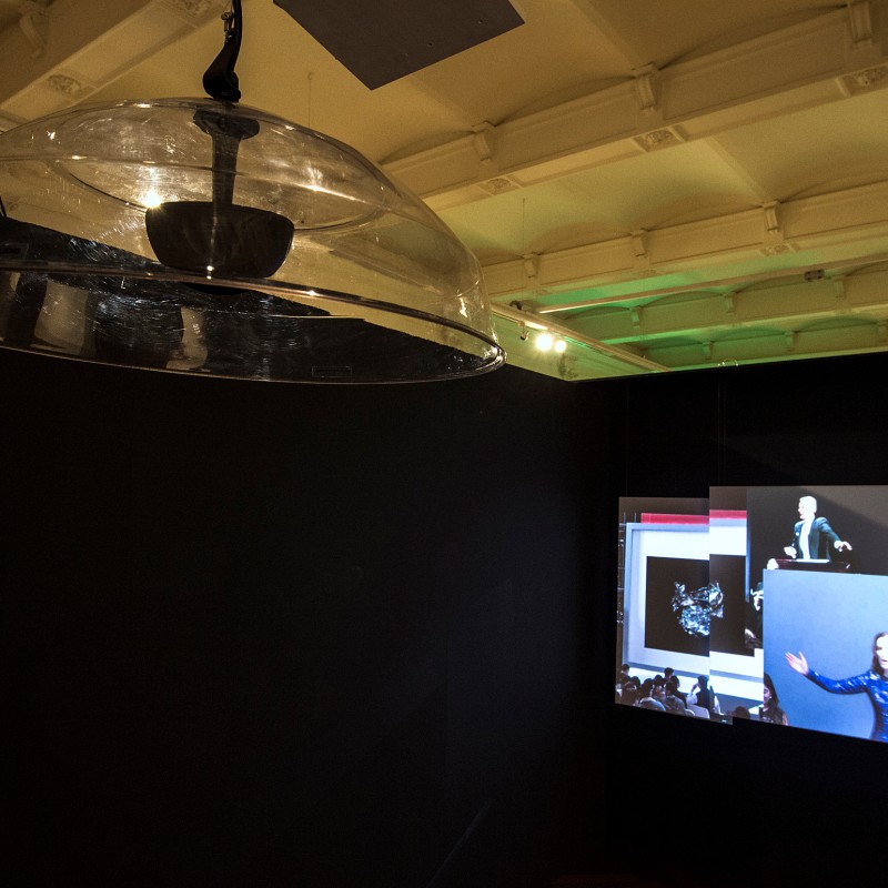 Large transparent dome hanging from the ceiling, projection with multiple overlapping images on a dark wall, including
                                 a person with outstretched arms. (AI-generated alt-text, generated with GPT-4.1-mini)