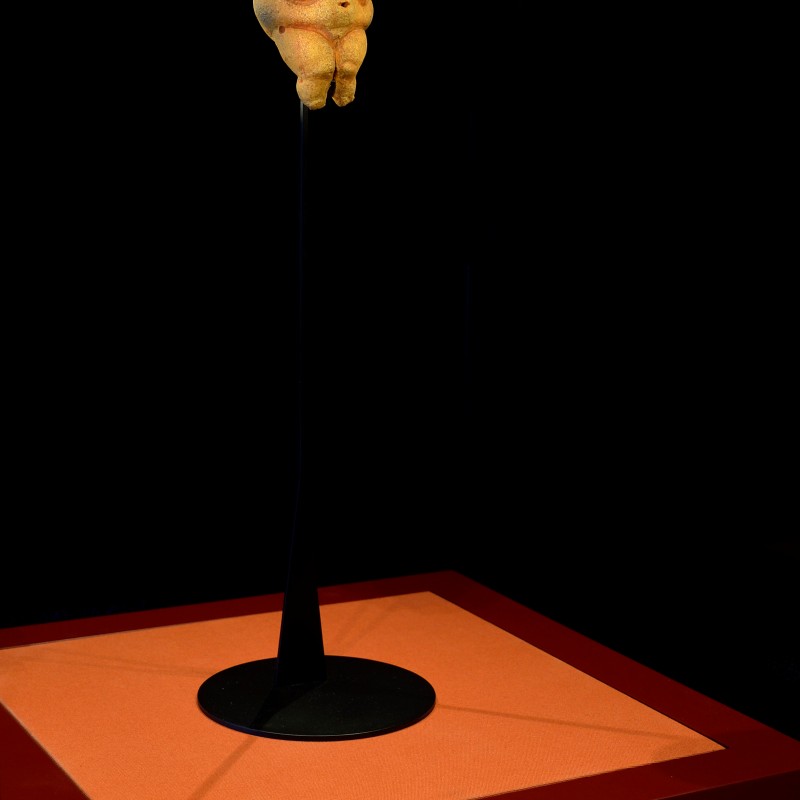 The Venus of Willendorf, a small prehistoric female figurine with pronounced curves, stands upright on a slender
                                 stand, dramatically lit against a dark background. (AI-generated alt-text, generated with GPT-4.1-mini)