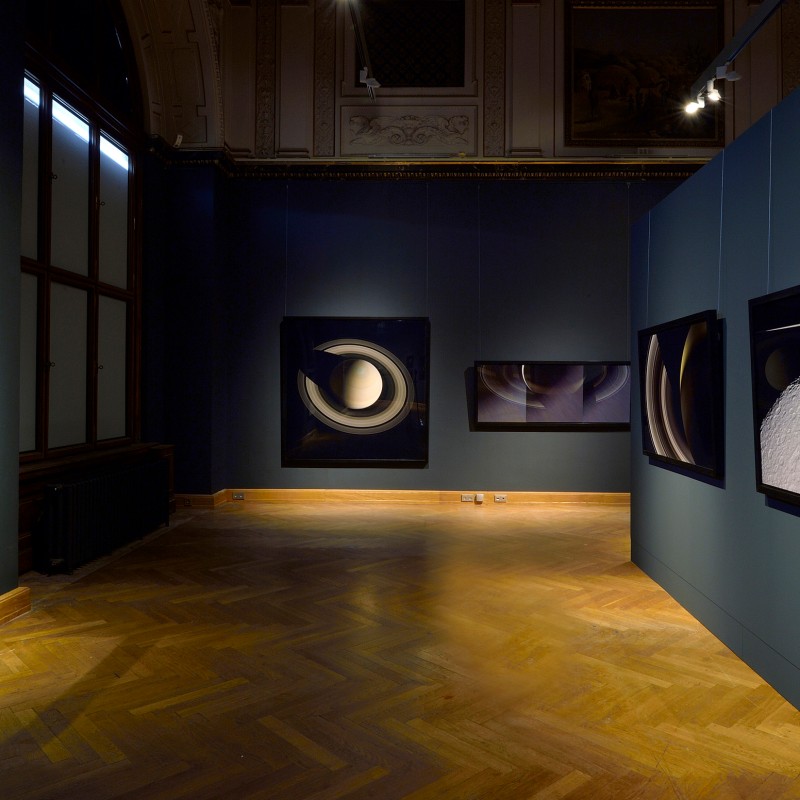 Dimly lit exhibition space featuring large photographs of planets and Saturn's rings on dark blue walls. (AI-generated
                                 alt-text, generated with GPT-4.1-mini)