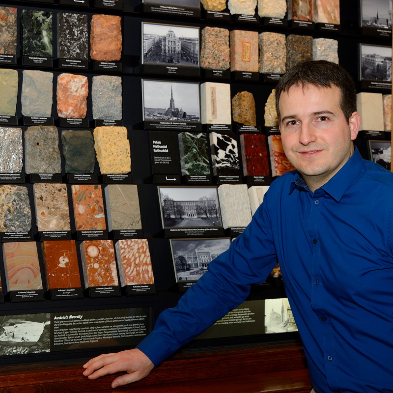 Man with short dark hair and blue shirt stands smiling in front of a wall with multiple rock samples. (AI-generated
                                 alt-text, generated with GPT-4.1-mini)