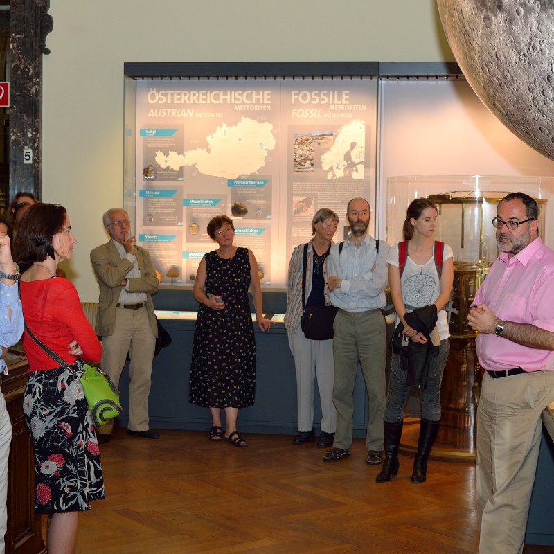 Group of adults stands in an exhibition room with large display
                                 boards about Austria, fossils, and lunar objects. A man with glasses and a pink shirt speaks, others listen. (AI-generated
                                 alt-text, generated with GPT-4.1-mini)