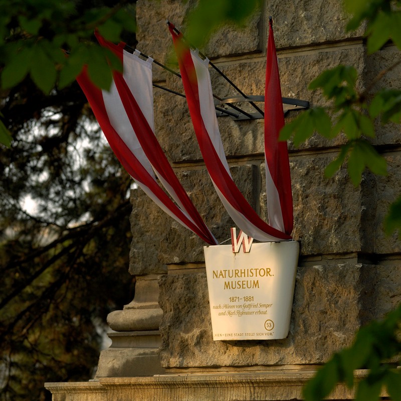 Red and white flags hang above a museum sign on a stone wall, surrounded by fresh green leaves. (AI-generated alt-text,
                                 generated with GPT-4.1-mini)