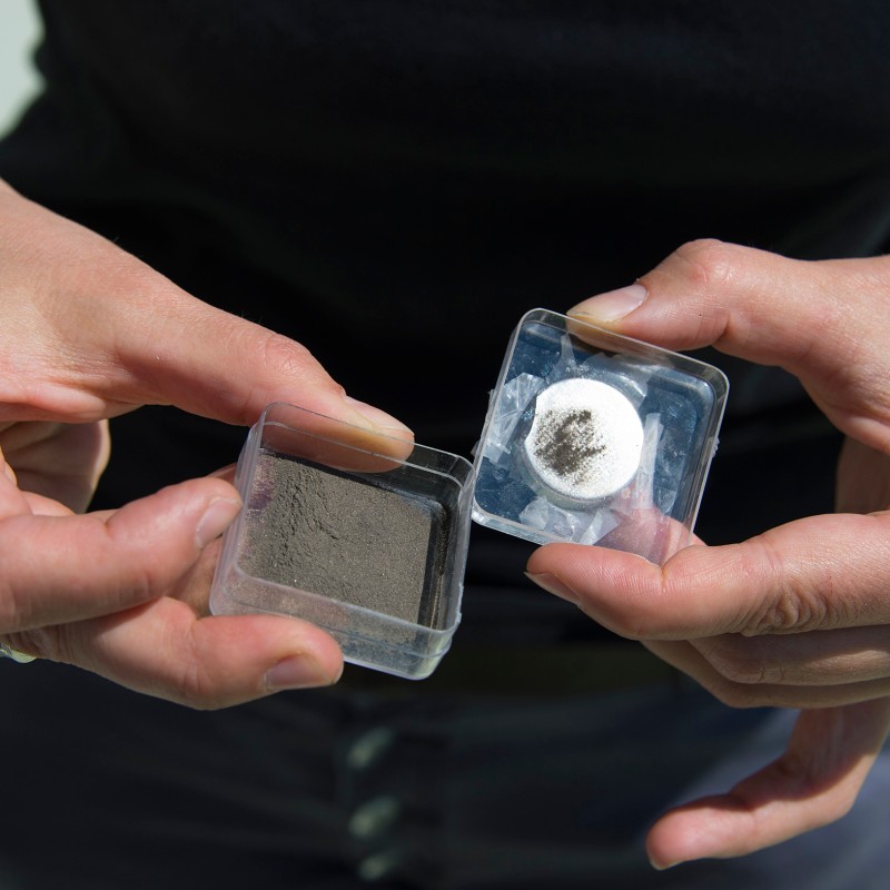 Person holding two small transparent containers, one with gray powder and one with a white disc showing a dark spot.
                                 (AI-generated alt-text, generated with GPT-4.1-mini)