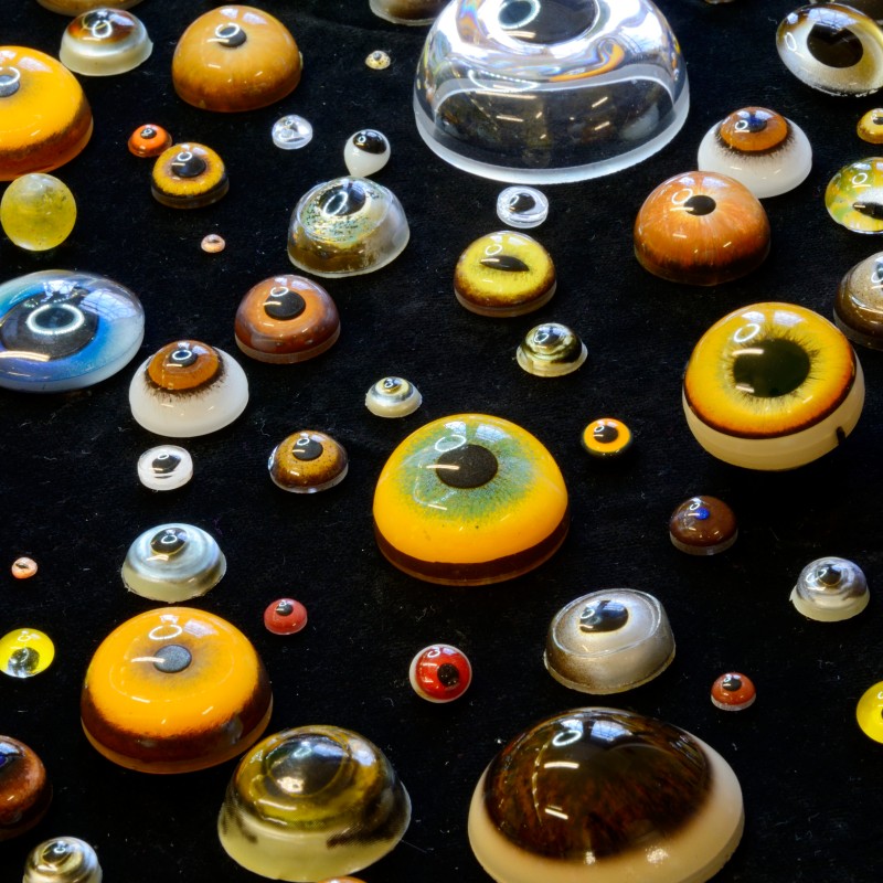 Bunte Glasaugen in verschiedenen Größen und Farben auf schwarzem Untergrund angeordnet. (KI-generierter Alt-Text,
                                 erstellt mit GPT-4.1-mini)