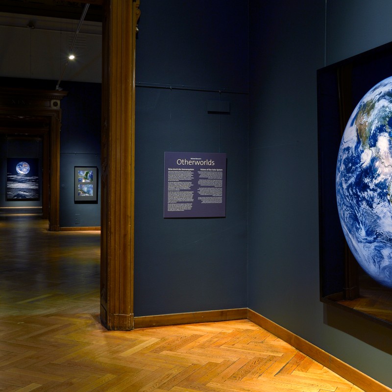 Large
                                 photograph of Earth from space, framing an exhibit with additional space images in a dimly lit museum hall. (AI-generated
                                 alt-text, generated with GPT-4.1-mini)