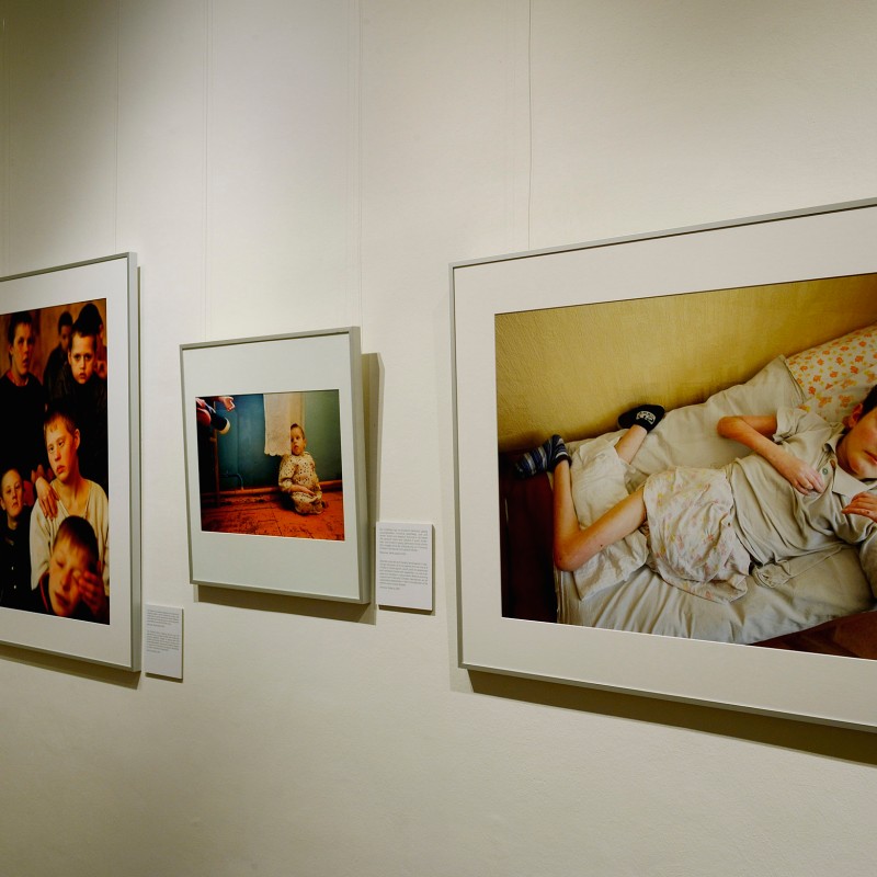 Exhibition view showing several framed photos on a white gallery wall, including a child lying on a bed wearing
                                 a light shirt with patterned bedding. (AI-generated alt-text, generated with GPT-4.1-mini)