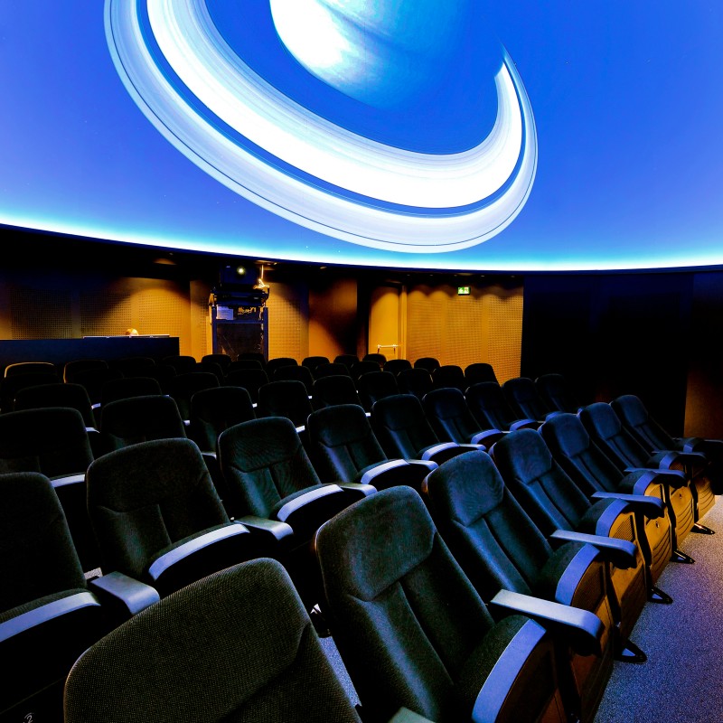 Auditorium with multiple rows of padded seats beneath a large projection of a ringed planet on the domed ceiling.
                                 (AI-generated alt-text, generated with GPT-4.1-mini)