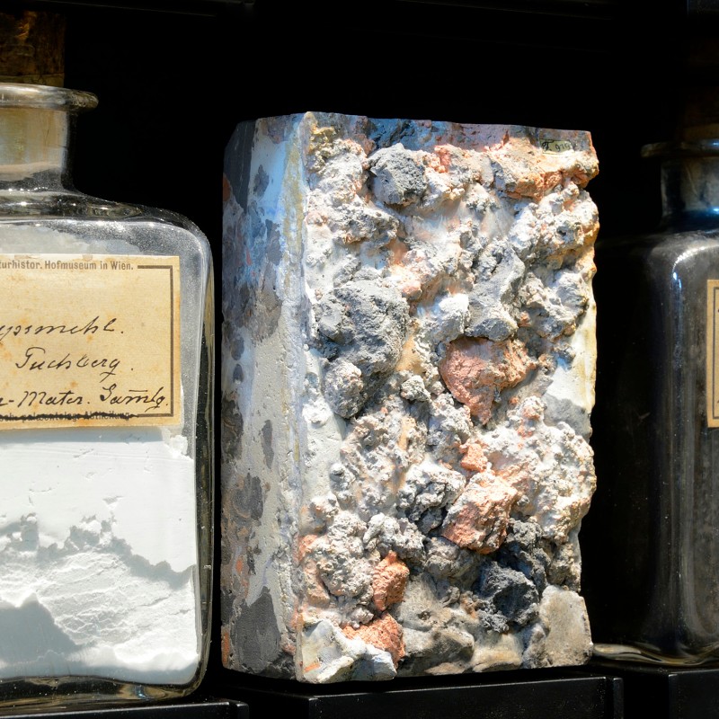 Glass containers with white and dark powdery samples and a rectangular mineral block with varied colors on a shelf.
                                 (AI-generated alt-text, generated with GPT-4.1-mini)