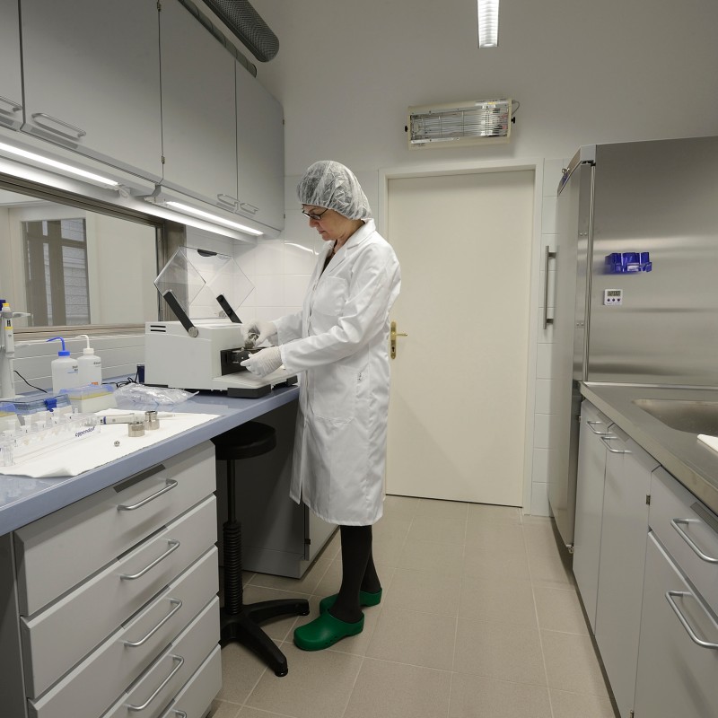 Laboratory room with a person in lab coat and hairnet standing at a white device. Surrounded by cabinets, work surfaces,
                                 and lab equipment. (AI-generated alt-text, generated with GPT-4.1-mini)
