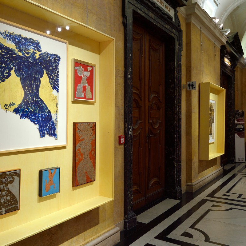 Exhibition wall with several framed collages and drawings in a museum hallway, illuminated by display lighting.
                                 (AI-generated alt-text, generated with GPT-4.1-mini)