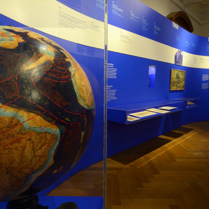 Room with a large globe in the foreground, marked with red lines; behind it, curved blue exhibition panels with
                                 text on the walls. (AI-generated alt-text, generated with GPT-4.1-mini)