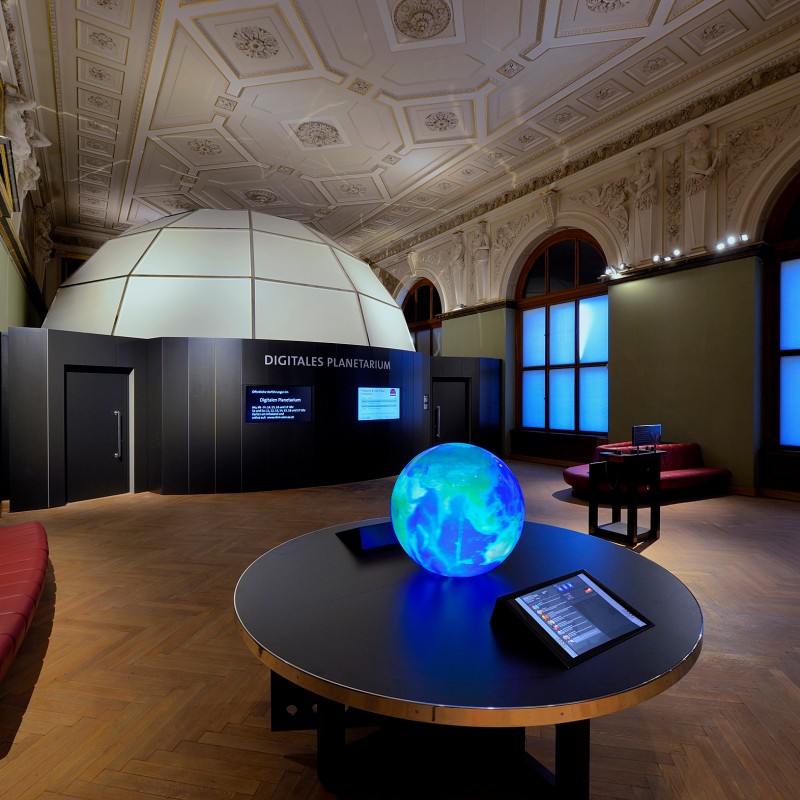 Moderner Raum mit Kuppelbau, leuchtender Kugel auf rundem Tisch, Informationsdisplay und großen Fenstern. (KI-generierter
                                 Alt-Text, erstellt mit GPT-4.1-mini)