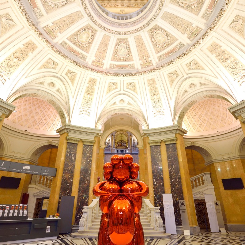 Large, shiny orange sculpture in the shape of a dog stands beneath an ornate domed ceiling in a grand museum hall.
                                 (AI-generated alt-text, generated with GPT-4.1-mini)