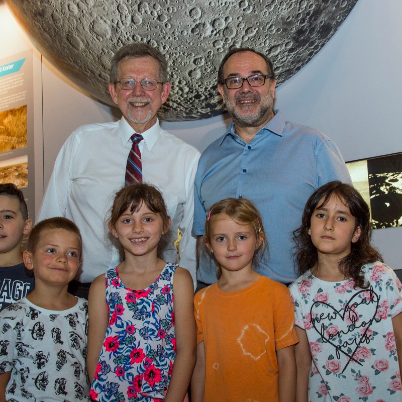 Two smiling men stand behind a group of four girls and two boys in front of a large moon
                                 illustration on an exhibition wall. (AI-generated alt-text, generated with GPT-4.1-mini)