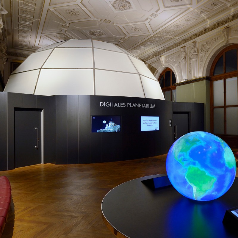 Large domed structure with the inscription 'Digitales Planetarium', in front a glowing digital globe on a round
                                 table. (AI-generated alt-text, generated with GPT-4.1-mini)