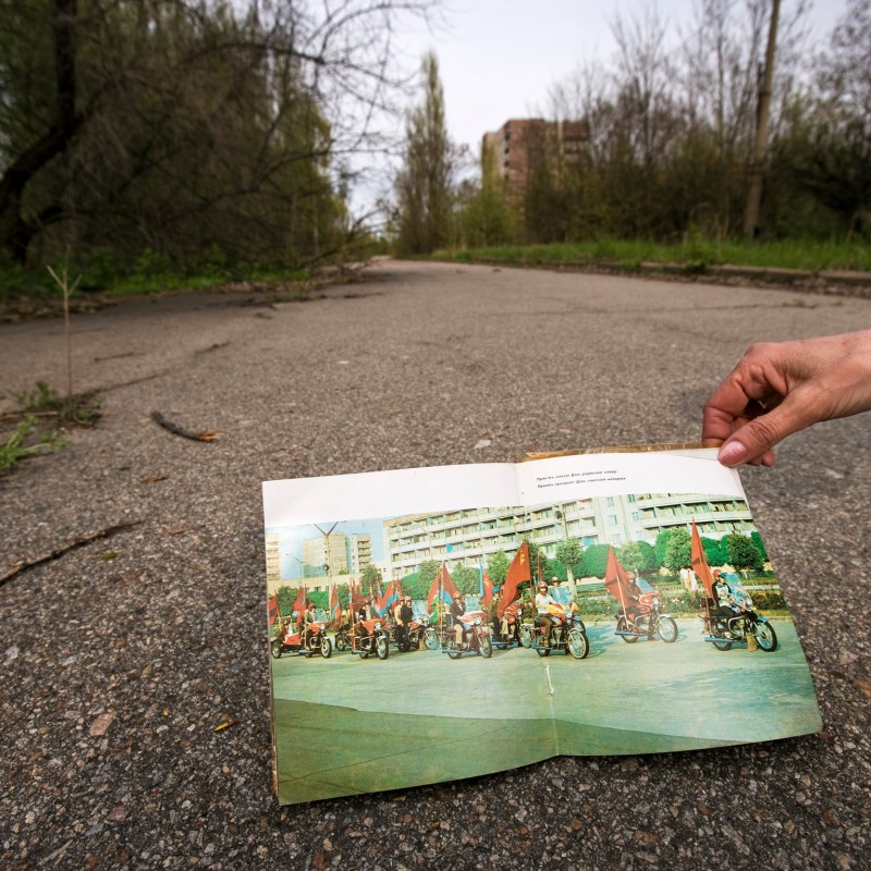 A hand holds an open photo of a parade with red flags on motorcycles on a street; abandoned, overgrown buildings
                                 in the background. (AI-generated alt-text, generated with GPT-4.1-mini)