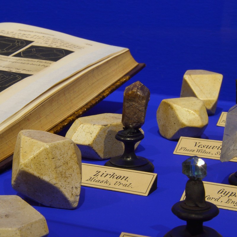 Various mineral and crystal models on black stands and as solid shapes beside an open book with crystal sketches
                                 (AI-generated alt-text, generated with GPT-4.1-mini)