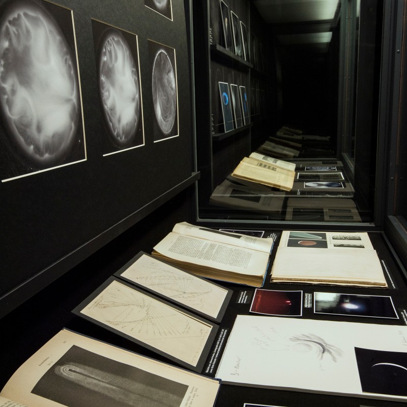 Several
                                 open books and panels with black-and-white and color astronomical images are arranged next to each other in a glass display
                                 case, with framed astronomical photographs on the wall above. (AI-generated alt-text, generated with GPT-4.1-mini)