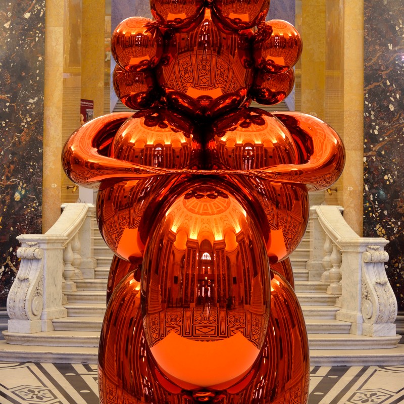 Large, shiny orange sculpture with rounded shapes in a historic, ornately decorated interior on a patterned floor.
                                 (AI-generated alt-text, generated with GPT-4.1-mini)