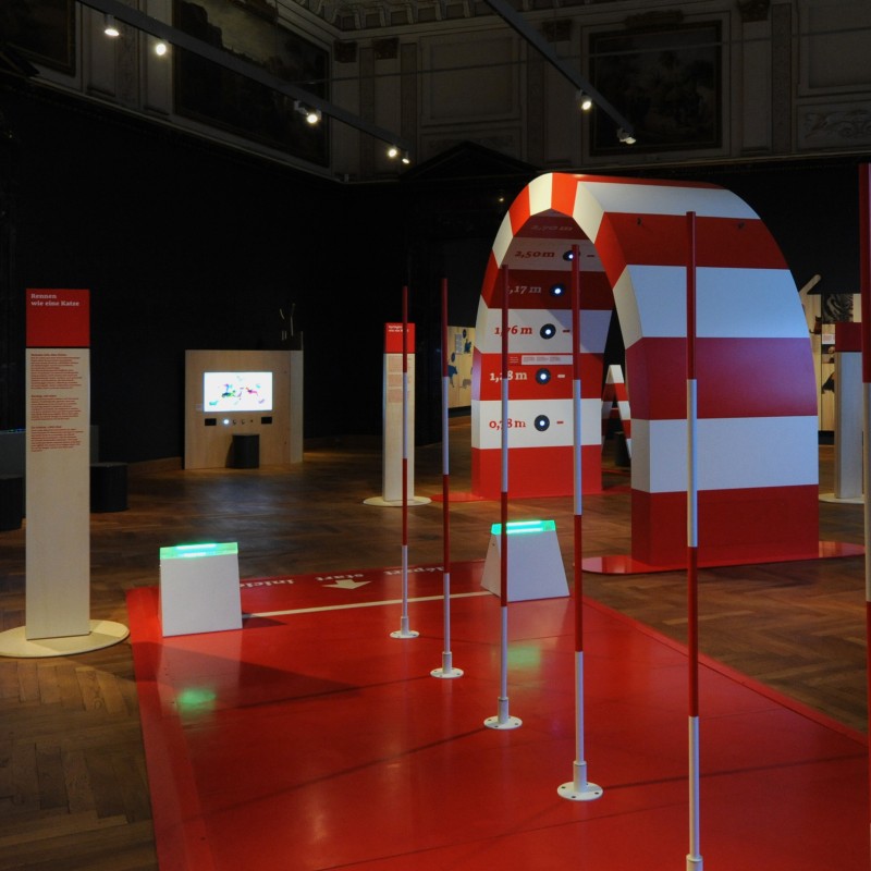 Red-and-white striped gateway with several pillars and digital screens in an exhibition room with wooden flooring.
                                 (AI-generated alt-text, generated with GPT-4.1-mini)
