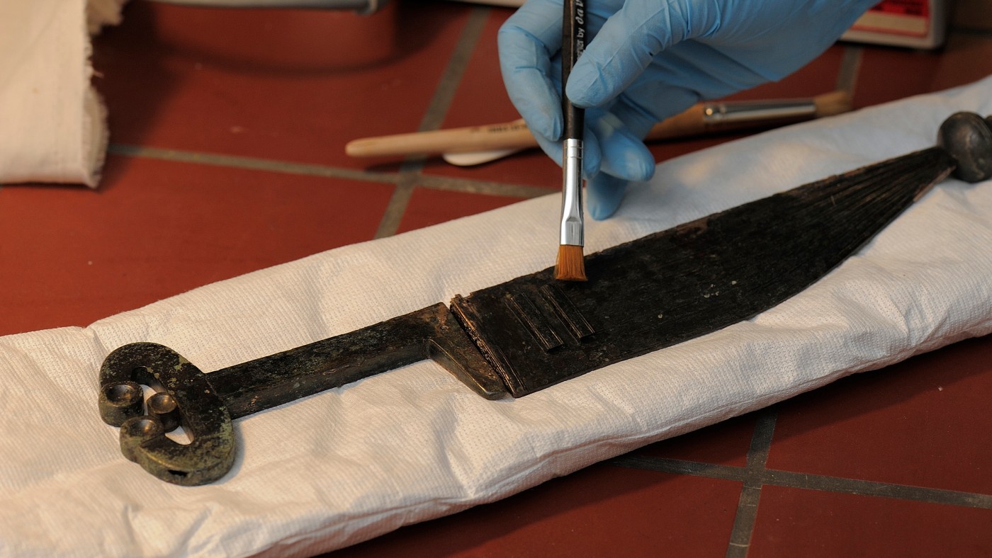 : Hand mit blauem Handschuh säubert eine antike Klinge mit einem Pinsel; die Klinge liegt auf weißem Polstermaterial. (KI-generierter Alt-Text, erstellt mit GPT-4.1-mini)