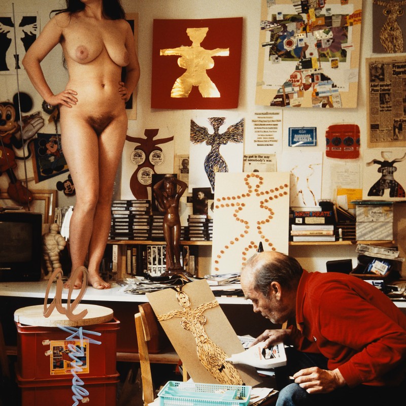 Nackte Frau mit langen dunklen
                                 Haaren in einem Atelier, umgeben von Kunstwerken mit weiblichen Figuren. Ein älterer Mann in rotem Pullover bearbeitet in
                                 einer sitzenden Position ein Kunstwerk. Wände sind voll mit Collagen und Drahtfiguren, Regale mit Kassetten und Büchern. (KI-generierter
                                 Alt-Text, erstellt mit GPT-4.1-mini)