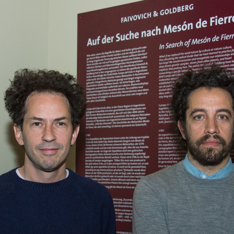 Two men with curly hair stand in front of an information board titled 'Auf der Suche nach Mesón de Fierro'. (AI-generated
                                 alt-text, generated with GPT-4.1-mini)