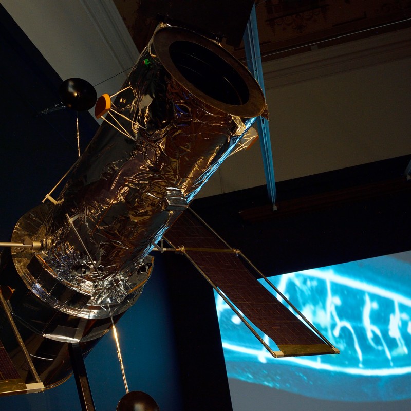 Modell
                                 des Weltraumteleskops Hubble mit reflektierender Oberfläche und ausgebreiteten Solarpaneelen vor einer projizierten Abbildung.
                                 (KI-generierter Alt-Text, erstellt mit GPT-4.1-mini)