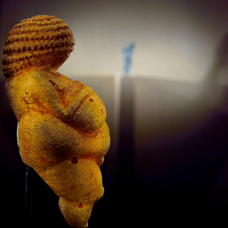 Close-up of the Venus of Willendorf, a small prehistoric female figurine with pronounced body features, illuminated
                                 with cast shadow on the wall. (AI-generated alt-text, generated with GPT-4.1-mini)