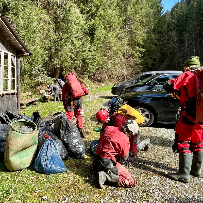 Höhlenforscher in roter Schutzkleidung und Stiefeln, mehrere Säcke mit Müll und Gerätschaften vor einer Holzhütte
                                 im Wald. (KI-generierter Alt-Text, erstellt mit GPT-4.1-mini)