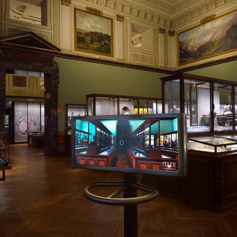 Ausstellungssaal
                                 mit Glasvitrinen, Holzfußboden, großen Gemälden an den Wänden und einem Bildschirm in der Mitte des Raums. (KI-generierter
                                 Alt-Text, erstellt mit GPT-4.1-mini)